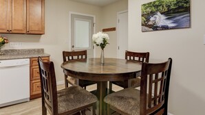Uma área de refeições com uma mesa de madeira redonda, quatro cadeiras, um vaso com flores brancas, armários de madeira, uma máquina de lavar louça e uma pintura de uma cascata na parede.