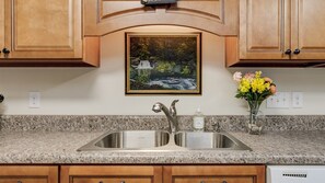 A kitchen with granite countertops, stainless steel double sink, wooden cabinets, a painting of a waterfall, and a vase with flowers.