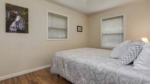 A bedroom with a bed, two windows with blinds, a framed picture on the wall, and a wooden floor.