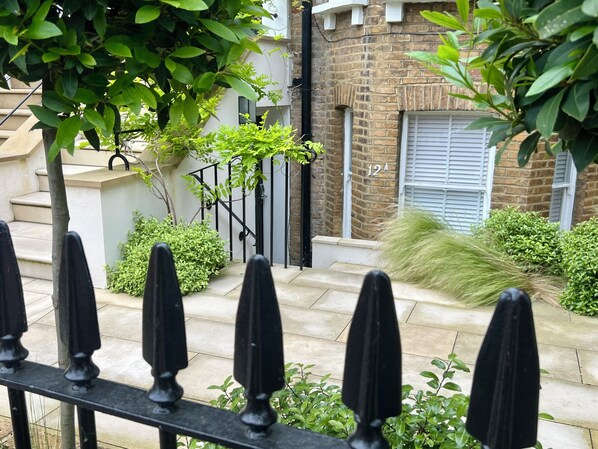 A residential street with a brick house, a black metal fence, and green foliage.