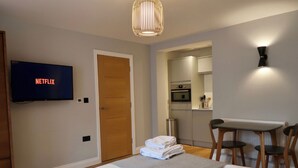 Ein Hotelzimmer mit Bett, einem Fernseher, der 'NETFLIX' zeigt, einer Holztür, einer Küchenzeile und einem kleinen Esstisch mit Stühlen.
