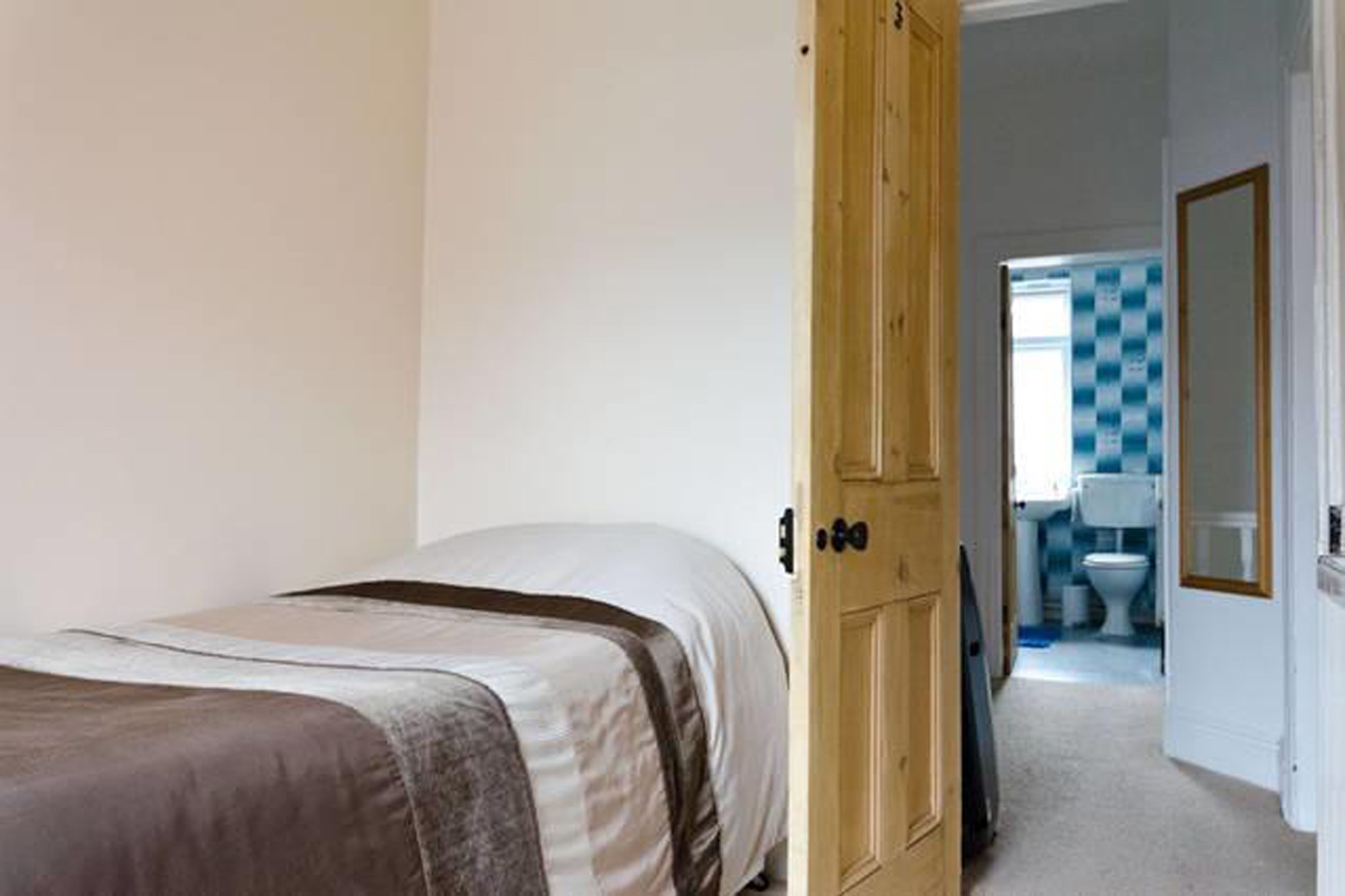 A bedroom with a bed, a wooden door, and a view into a bathroom with a checkered shower curtain.