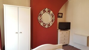 Un dormitorio con un armario blanco, un espejo de marco ornamentado, un televisor sobre una cómoda y una cama con colcha a rayas.