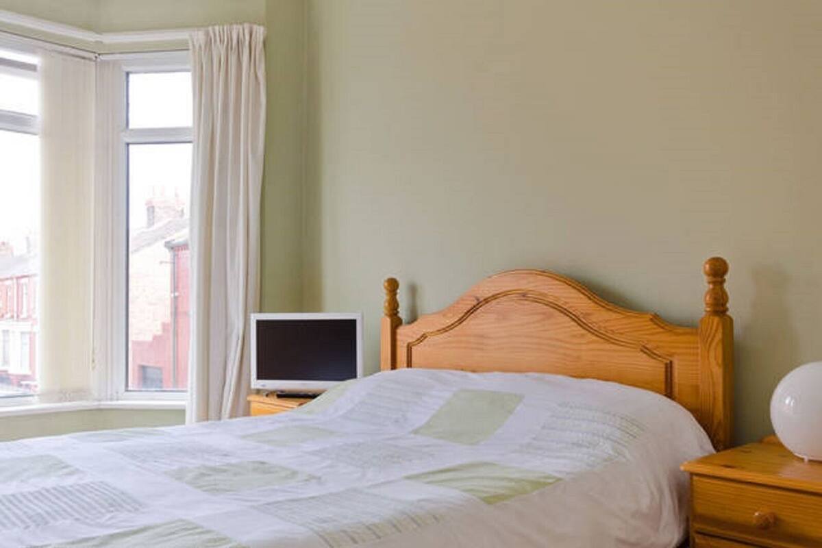 A bedroom with a wooden bed, a bedside table with a TV, and a window with curtains.
