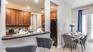 A modern kitchen with wooden cabinets, a black refrigerator, and a dining area with grey chairs and a table set for two. - Stylish NEW Renovation in Reunion Resort - Large Patio and Pool Deck 3bd/3.5ba #3ru456 (Kissimmee)