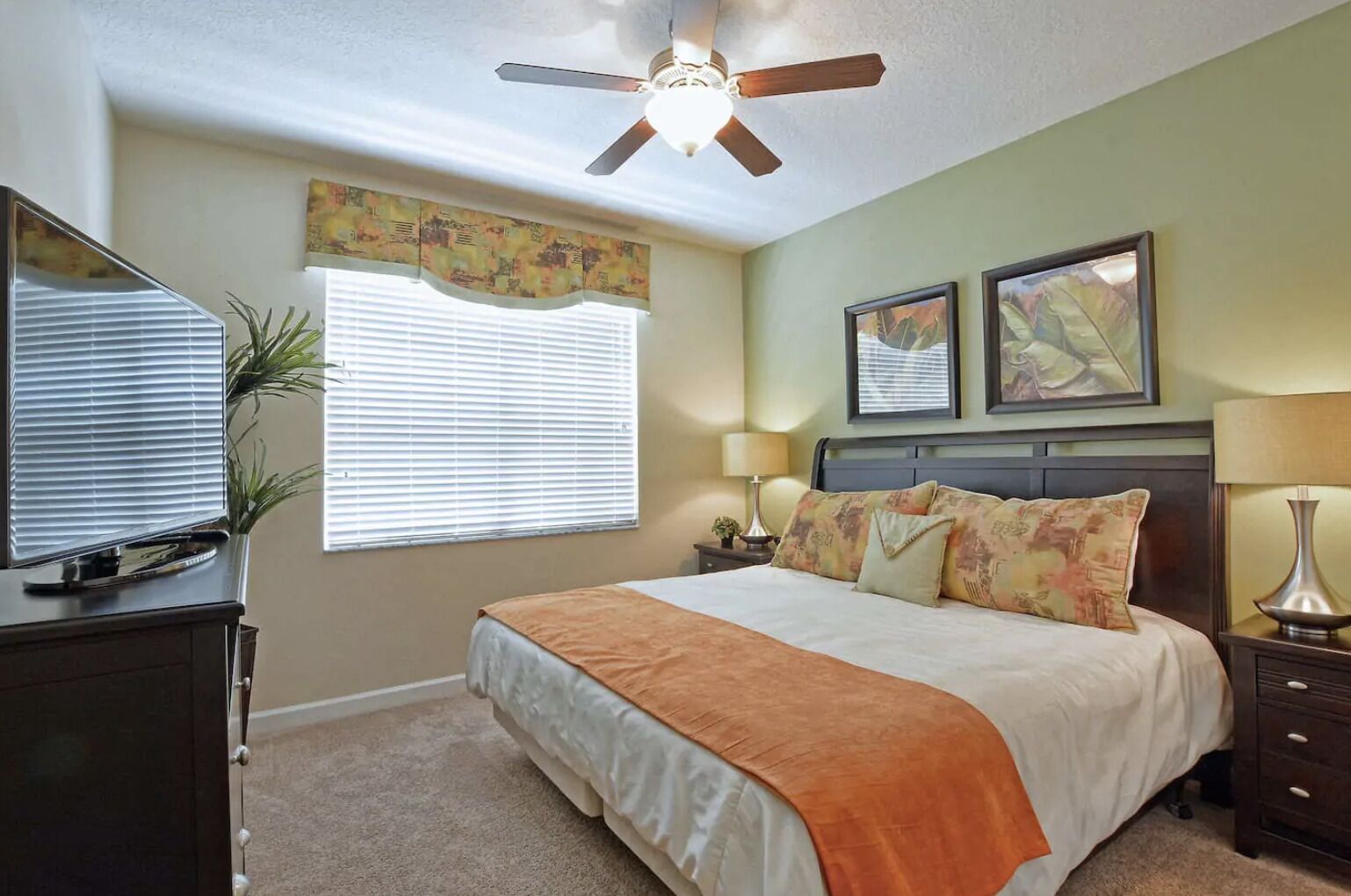 A bedroom with a bed, a ceiling fan, a television, a window with blinds, and two framed pictures on the wall.