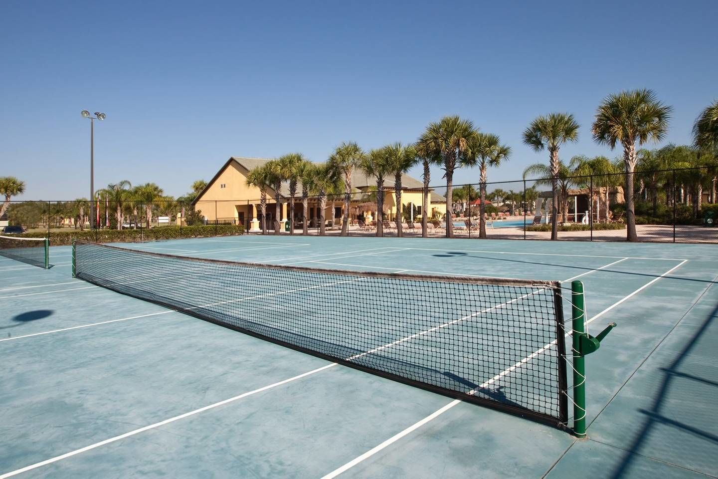 An outdoor tennis court with a net, surrounded by palm trees and a building in the background.