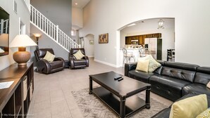 Ein geräumiges Wohnzimmer mit einer schwarzen Ledersofa, einem Couchtisch und zwei Ledersesseln. Eine Treppe führt zu einer oberen Ebene.
