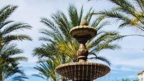 A fountain with multiple tiers and water spouts, surrounded by palm trees and a building with a tiled roof in the background. - Stylish and Accessible in Vista Cay With Lake View - 3bd/2ba Condo 3vc114 (Orlando)