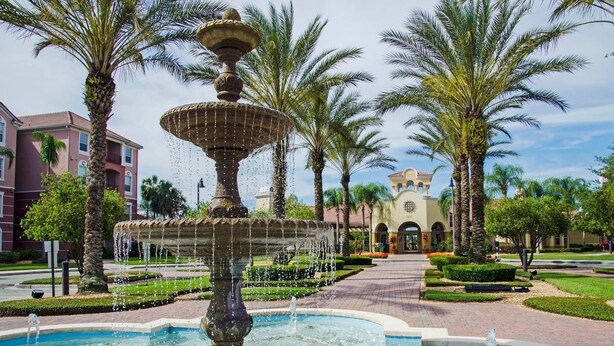 A fountain with multiple tiers and water spouts, surrounded by palm trees and a building with a tiled roof in the background. - Stylish and Accessible in Vista Cay With Lake View - 3bd/2ba Condo 3vc114 (Orlando)