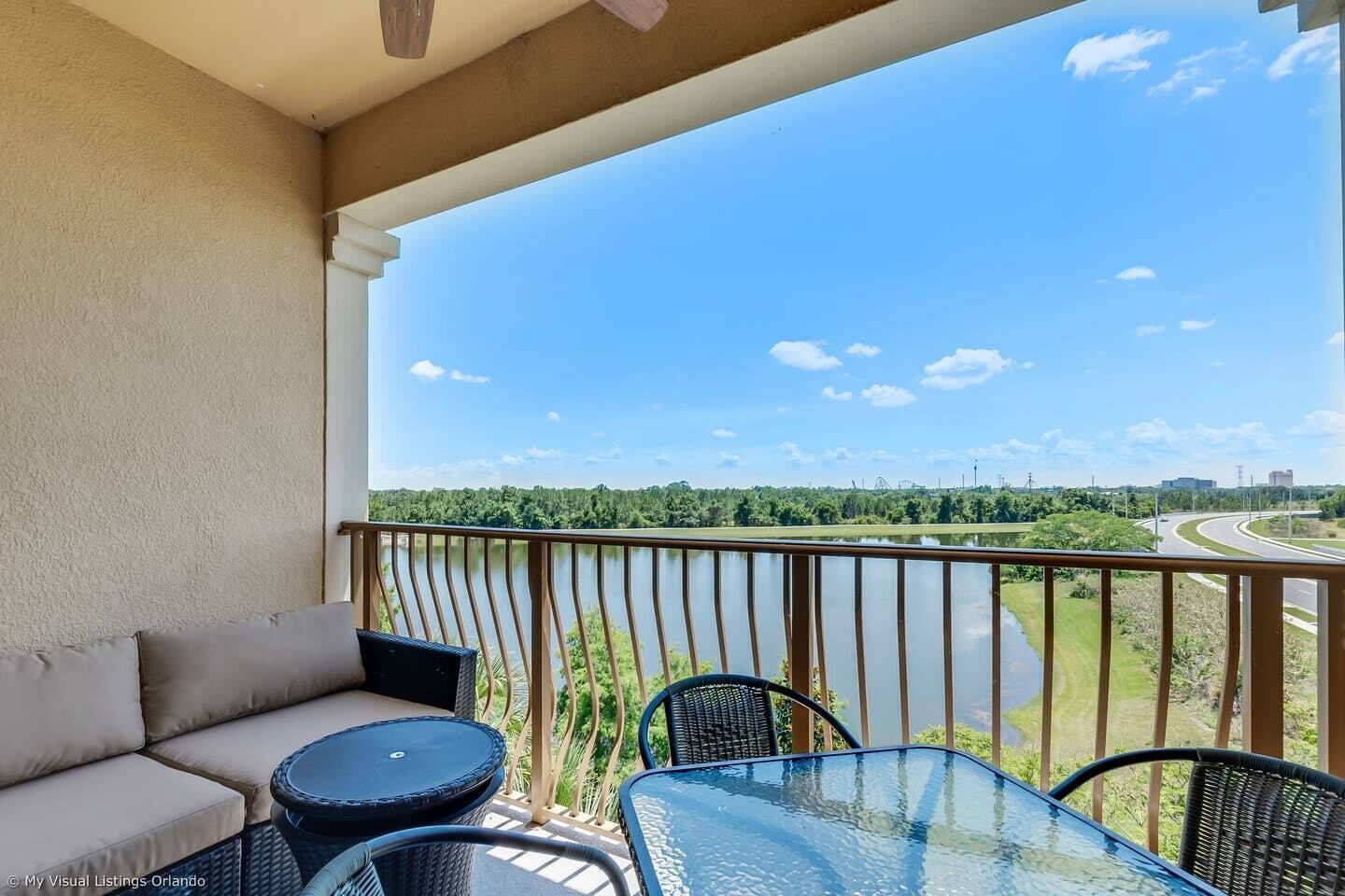 A balcony with a sofa and two round tables, overlooking a lake and a highway.