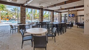 An outdoor dining area with tables and chairs, a pool with palm trees, and a stone wall with a television.