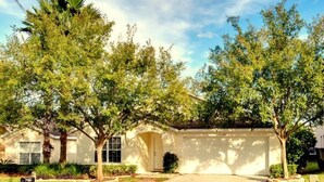 A single-story house with a front garage door, surrounded by mature trees and a well-maintained lawn. - Comfy and Spacious Near Disney With Pool - 4bd/3ba #4hl124 (Davenport)