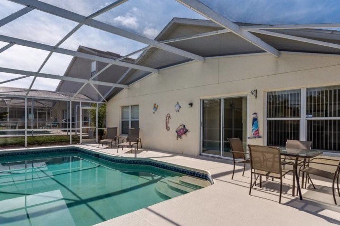 A covered outdoor swimming pool area with a glass roof, a patio with chairs, and a sliding glass door leading to a covered patio area.