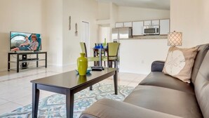 A living room with a sofa, a coffee table, and a TV. There is a kitchen in the background with cabinets and a microwave. - Comfy and Spacious Near Disney With Pool - 4bd/3ba #4hl124 (Davenport)