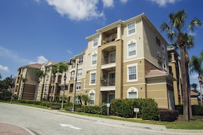 A multi-story residential building with balconies, a landscaped front yard, and palm trees.
