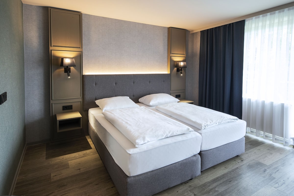 Une chambre d’hôtel moderne équipée d’un lit double, de tables de chevet, de lampes murales et d’une grande fenêtre avec des rideaux.