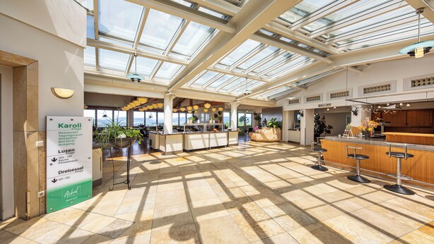 A spacious indoor area with a high ceiling and skylights, featuring a bar area with stools and a reception desk. - Michel Hotel Karoli Waldkirchen (Waldkirchen)