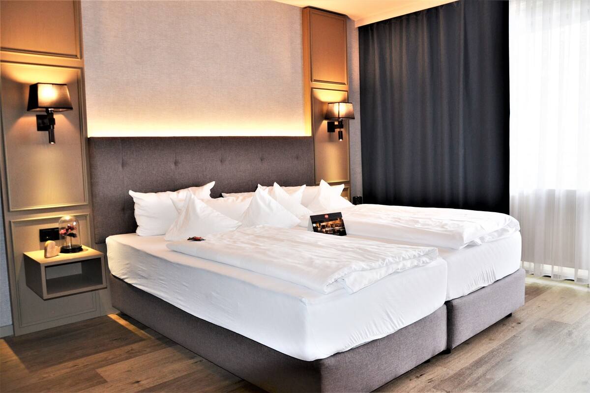Un lit double avec du linge de lit blanc, deux tables de chevet avec des lampes et une vue sur un rideau sombre.
