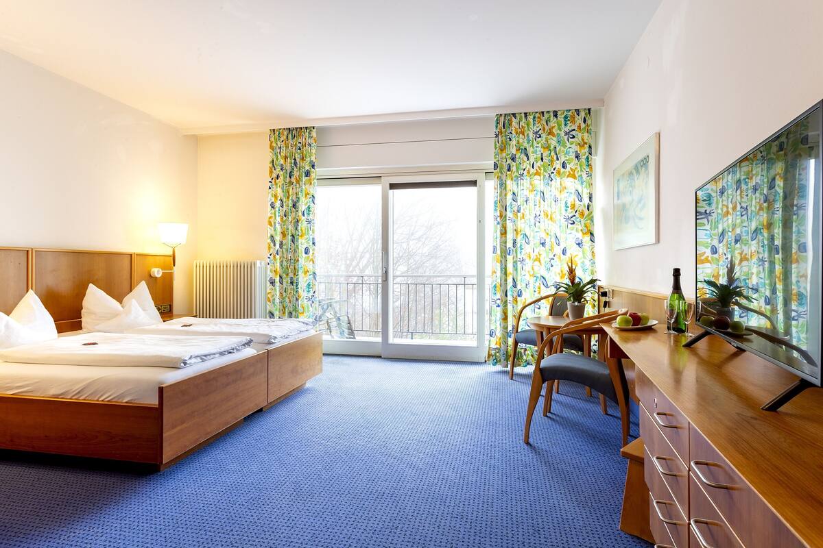 Une chambre d’hôtel avec un grand lit, un bureau avec une chaise, une télévision et un balcon avec des rideaux.