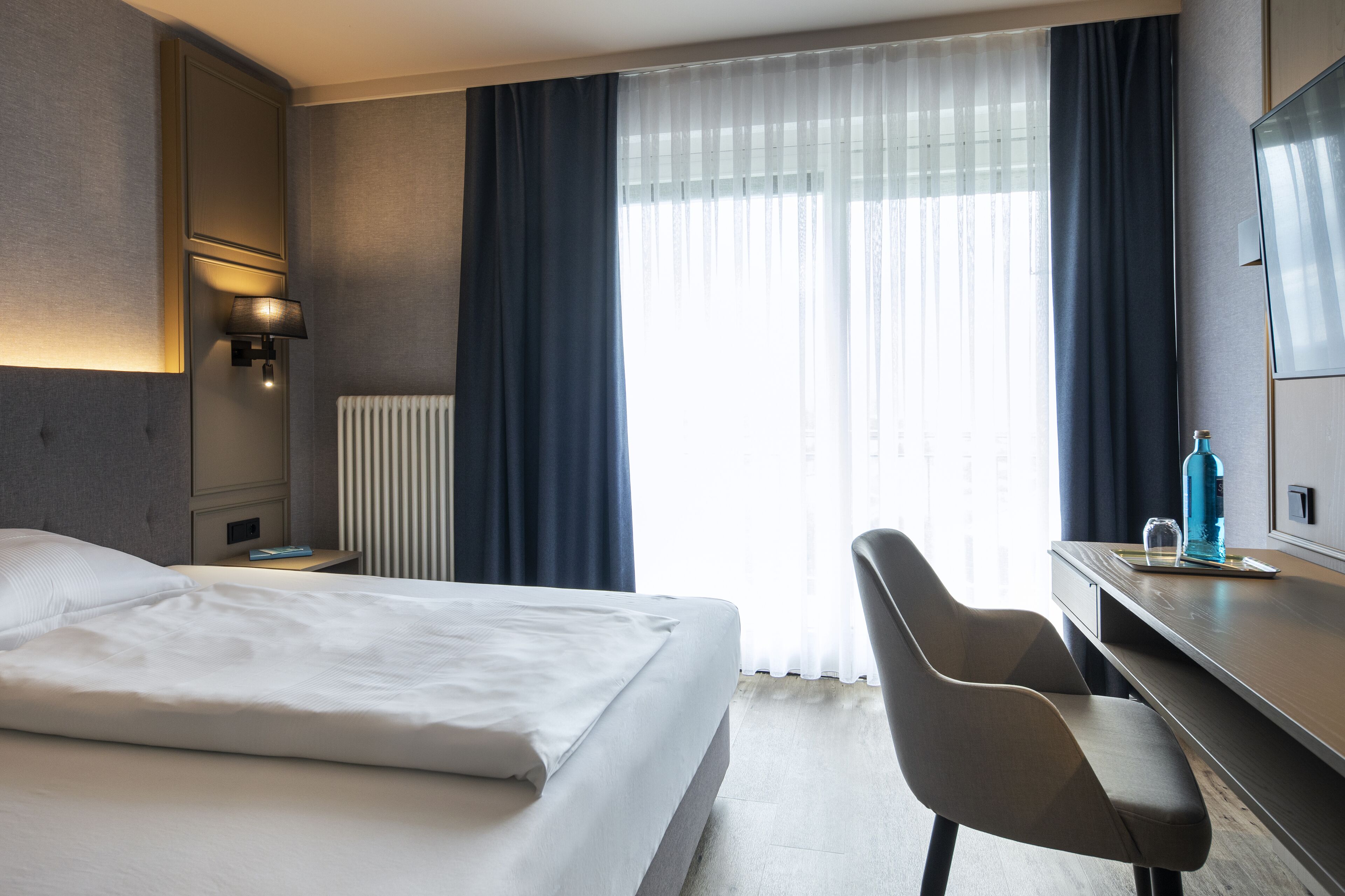 Habitación de hotel con cama, escritorio, silla, lámpara y ventana con cortinas.