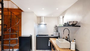 Une cuisine compacte équipée d’une cuisinière intégrée, d’un four à micro-ondes et d’un évier. Un escalier en bois se trouve à gauche, et une étagère contenant des ustensiles de cuisine est placée au-dessus de l’évier.