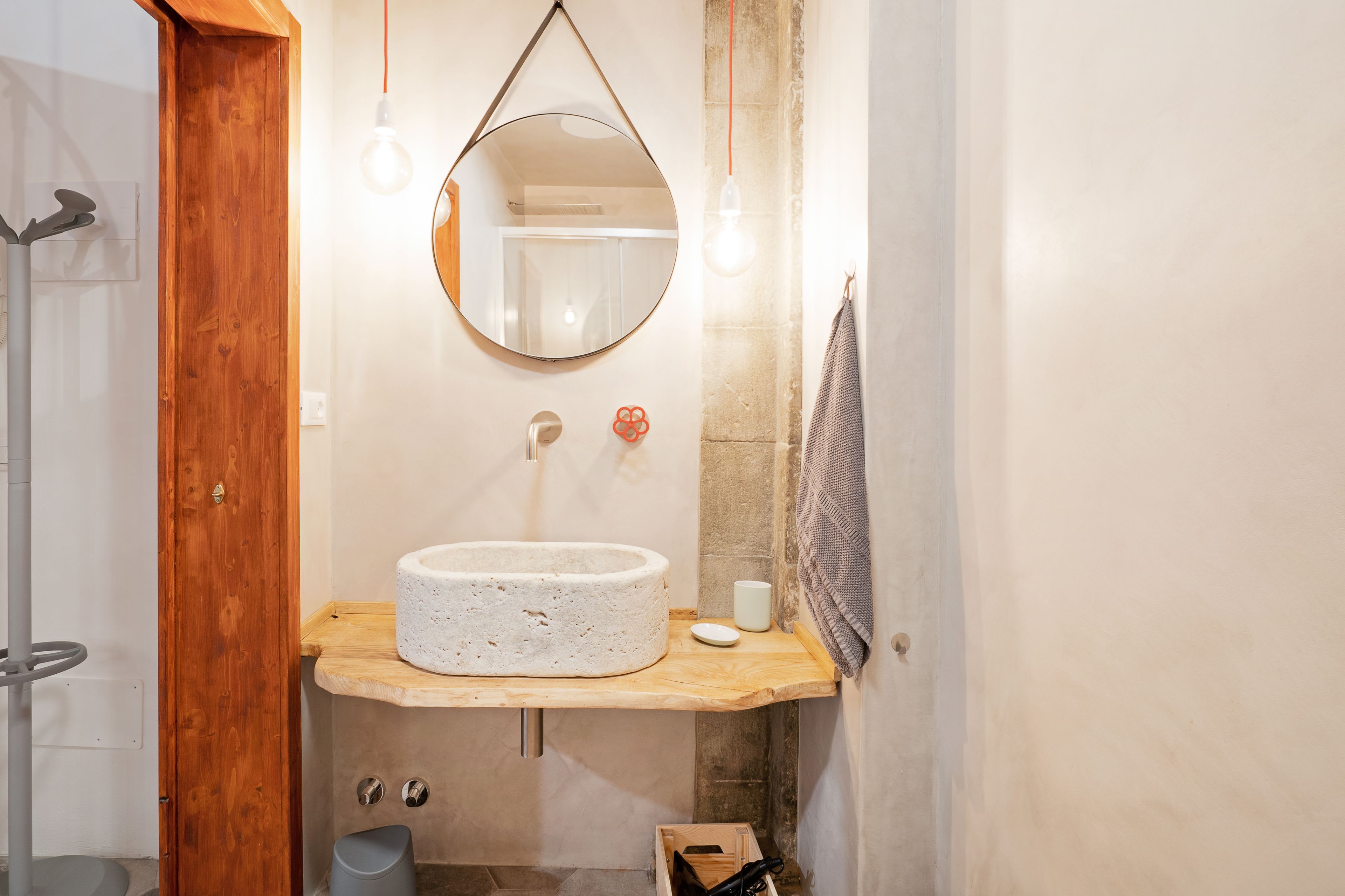 Un bagno con un lavandino rotondo e strutturato, un mobile contenitore in legno, uno specchio e un portascopevoli.