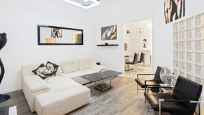 Ein modernes Wohnzimmer mit einer Couch, einem Couchtisch und einem schwarzen Sessel. An der Wand hängt ein Kunstwerk und eine Uhr.