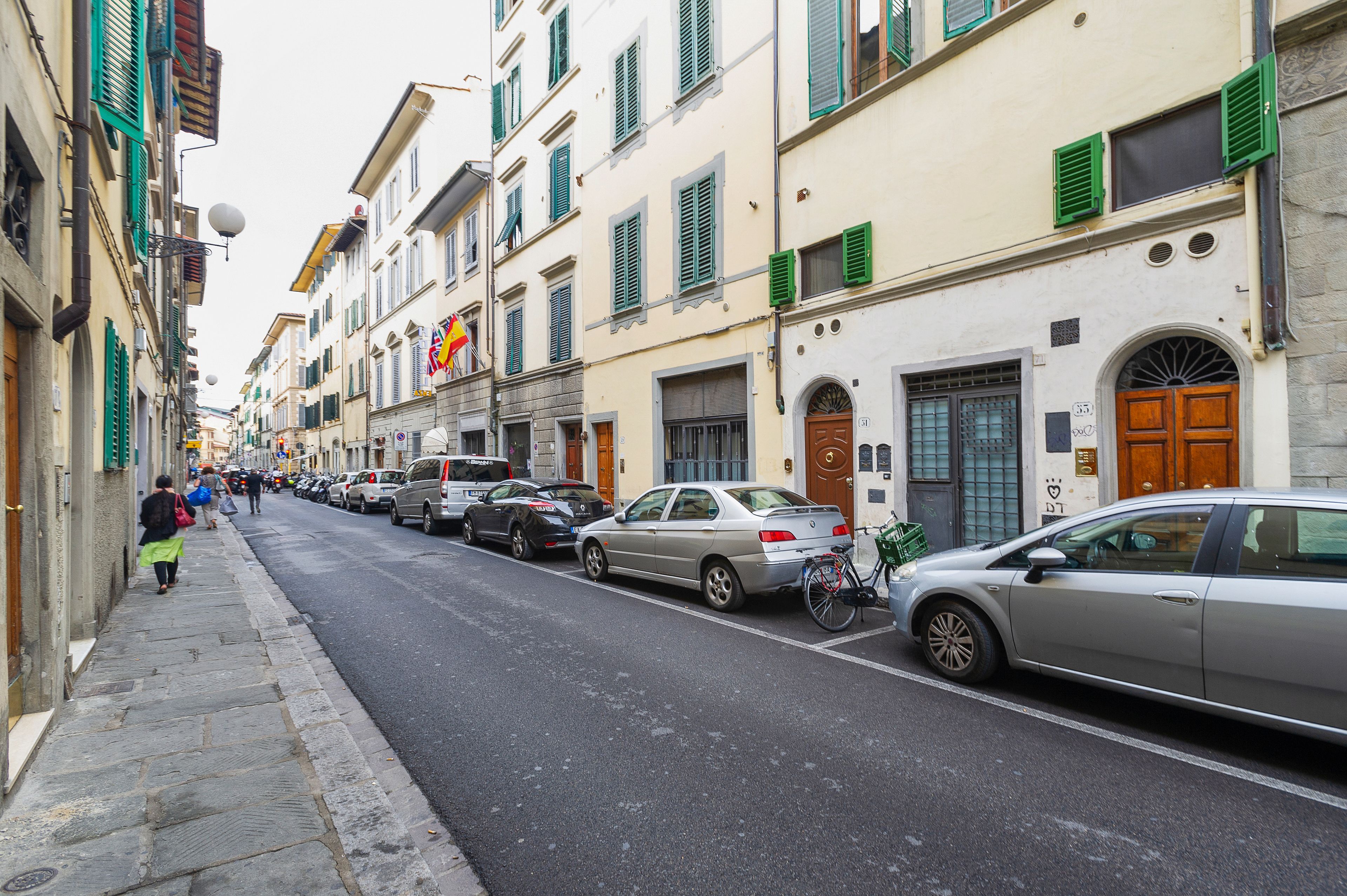Une rue bordée de voitures garées et d’immeubles aux volets verts.