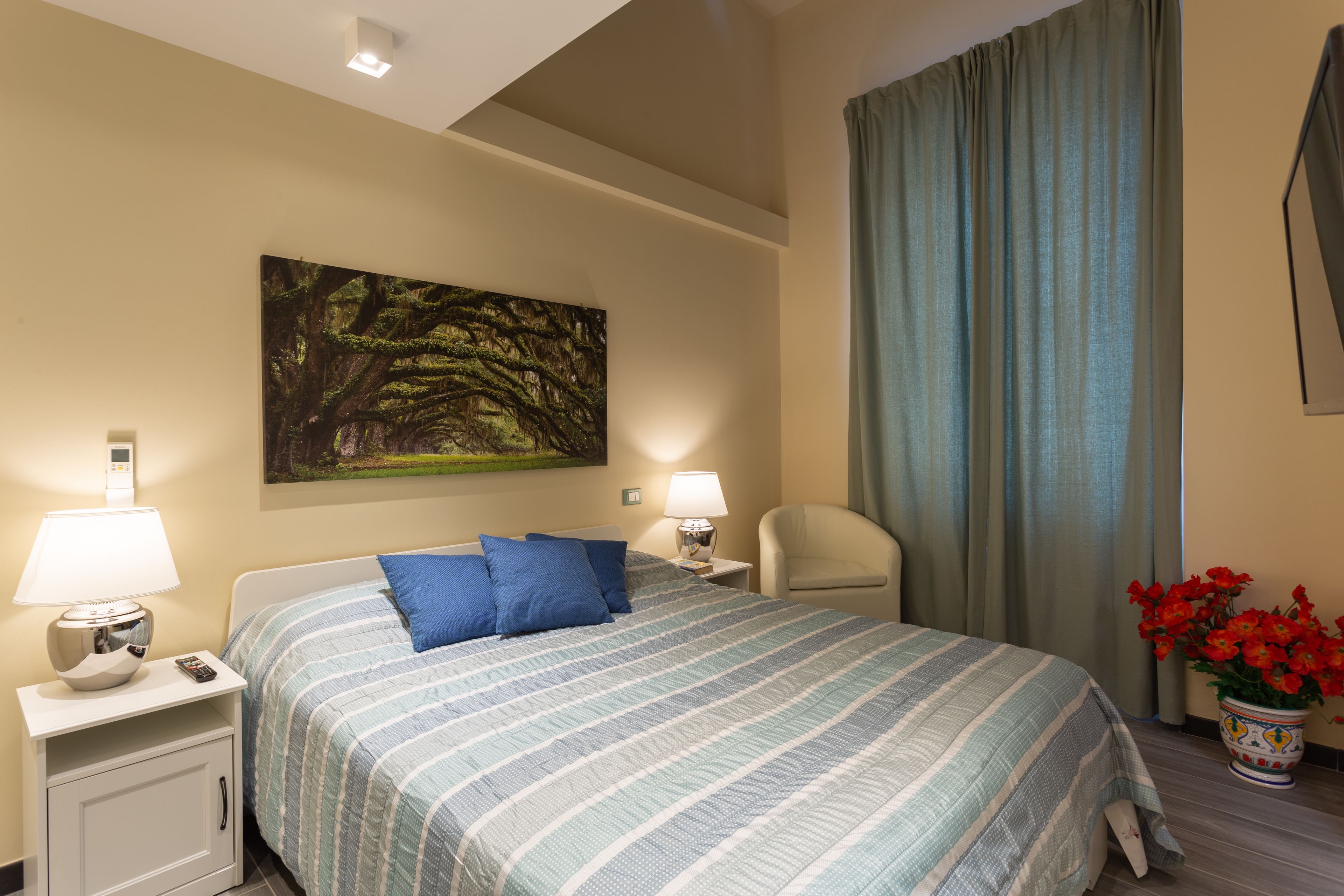 A hotel room with a bed, two bedside tables with lamps, a chair, a painting on the wall, and a potted plant.