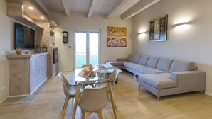 A modern living room with a glass dining table, a grey sofa, and a kitchen area with a TV and clock. - Accommodation Spaccasole 2 on Cortona (Foiano della Chiana)