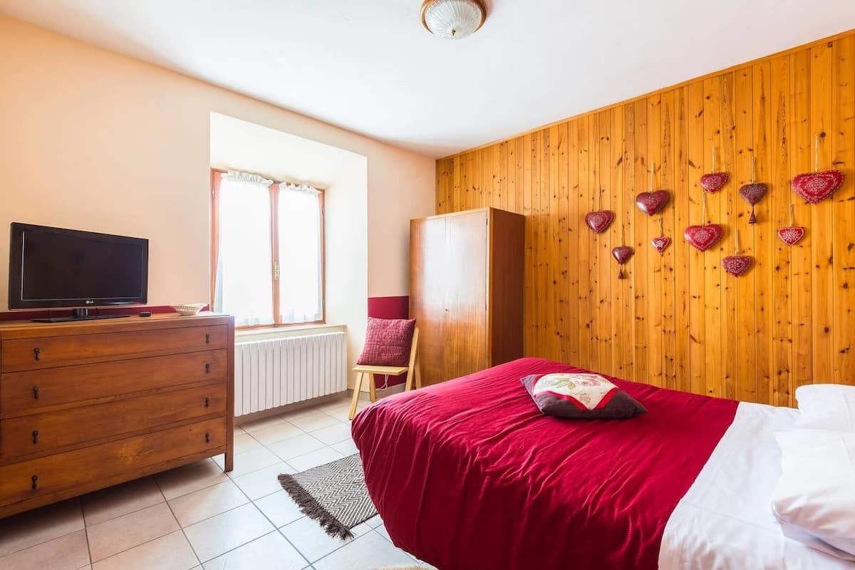 Un dormitorio con pared de madera, cama con ropa de cama roja, cómoda, televisor y ventana con cortinas.