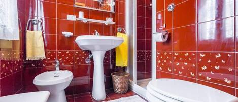 A bathroom with a white sink, bidet, and toilet, red tiled walls, and a white rug.