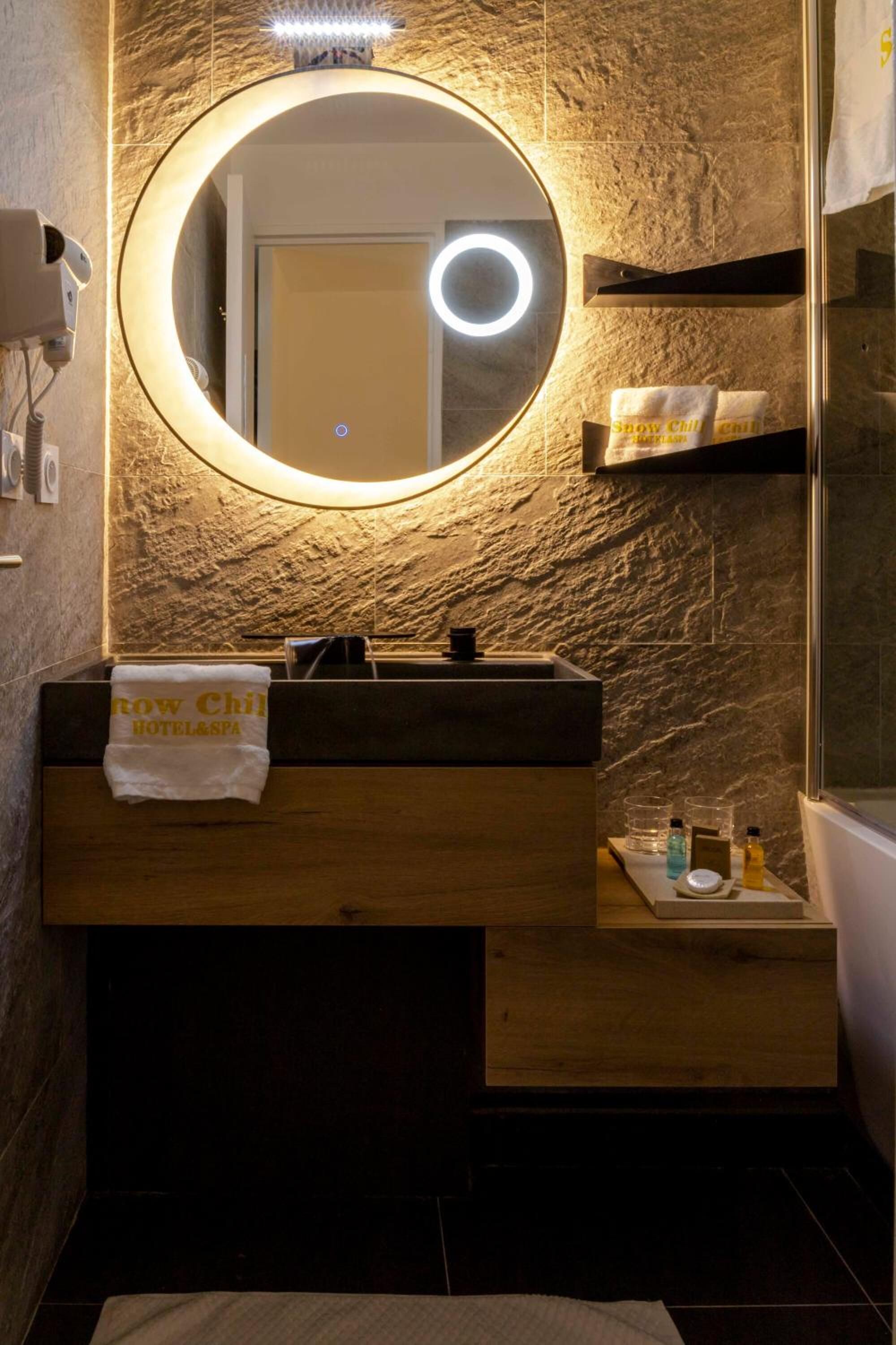 Un baño con un espejo redondo, un tocador de madera y un perchero con toalla.