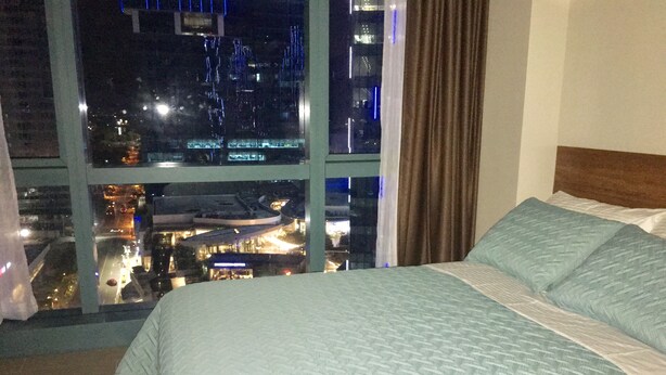 A hotel room with a large bed, a view of a cityscape through a window, and a curtain on the window. - BGC One Uptown 1 Bedroom Corner Unit 16th floor with City View (Taguig)