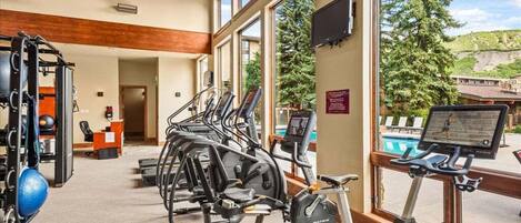 A gym with various exercise equipment, a reception desk, and large windows offering a view of a pool and trees.