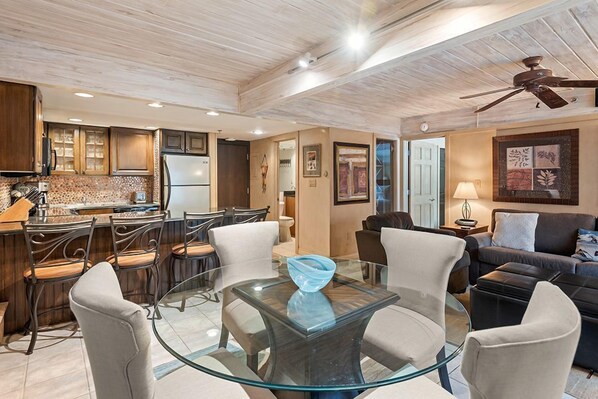 A modern kitchen with a dining area, featuring a glass table and chairs, wooden cabinets, and a ceiling fan.