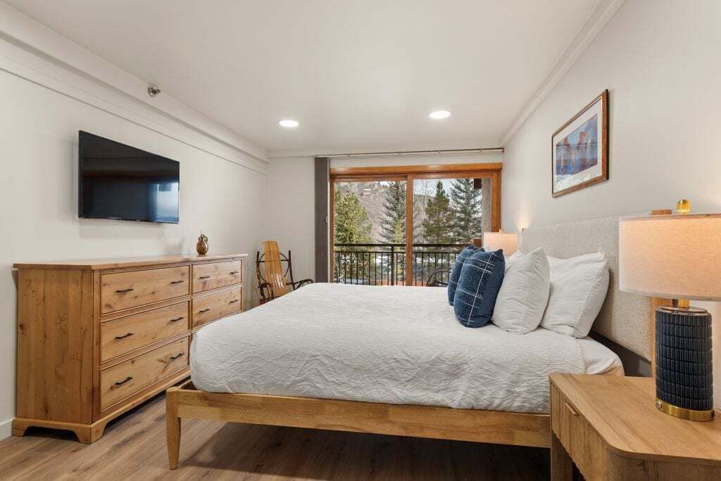 A bedroom with a wooden bed, a TV on the wall, a balcony with a view of trees, and a wooden dresser.