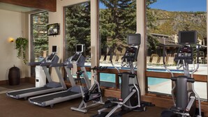 Gimnasio con cintas de correr, elípticas y una bicicleta estática, con vistas a una piscina y árboles.