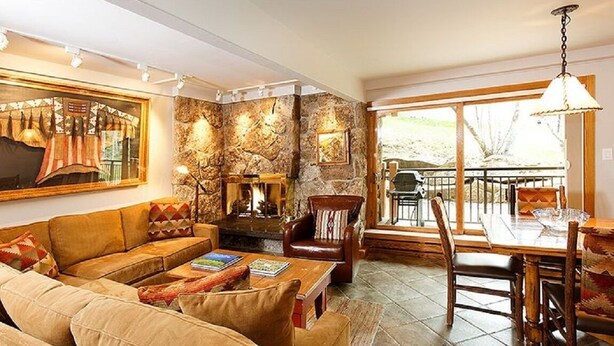 A cozy living room with a stone wall, a fireplace, and a dining area with a view. - Stonebridge Condos 707 1 Bdrm Slopeside (Snowmass Village)