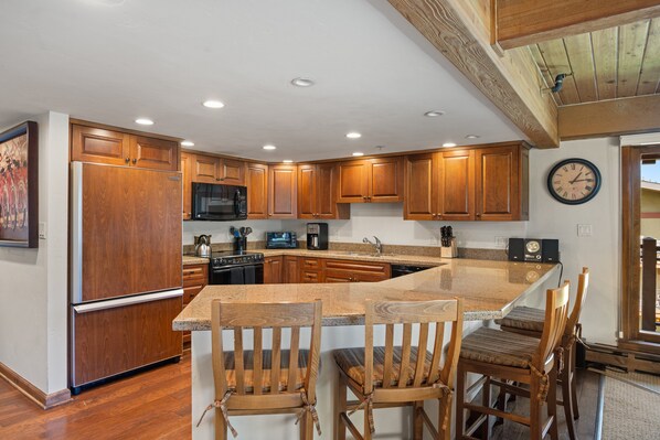A kitchen with wooden cabinets, a refrigerator, a microwave, and a sink. There are two wooden chairs with cushions. - Right on the Slopes Stonebridge Condo 610 2 Bedroom Den 3 BA (Snowmass Village)