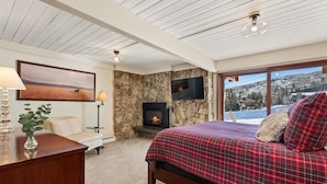 A bedroom with a stone fireplace, a bed with a plaid bedspread, a desk with a lamp, and a large window with a view of a snowy landscape.