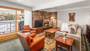 Uma sala de estar com parede de pedra, lareira, televisão e varanda com vista para uma cidade.