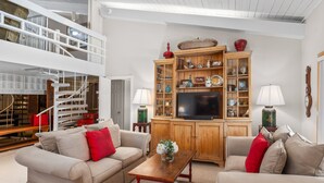 A spacious living room with a high ceiling, a wooden cabinet with glass doors, a television, and a staircase leading to an upper level. - Stonebridge Condo 729, 1 Bdrm and Loft, W/D, Slopeside (Snowmass Village)