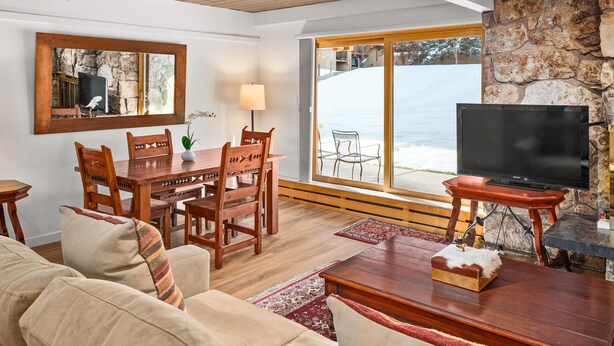 Un salon chaleureux avec un plafond en bois, une cheminée en pierre, un coin repas avec des meubles en bois et une grande fenêtre donnant sur l’extérieur.
