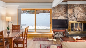 Un salon chaleureux avec une cheminée en pierre, des meubles en bois et une vue sur un paysage enneigé, visible à travers de grandes portes vitrées.