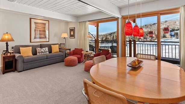 Ein geräumiges Wohnzimmer mit grauem Sofa, roten Sesseln, einem runden Holztisch und einem großen Fenster mit Blick auf einen Berg und Seilbahnen.