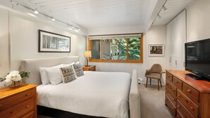 Ein Schlafzimmer mit Bett, Holzkommode, Fernseher und einem Fenster mit Blick auf Grünfläche.