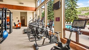Ein Fitnessstudio mit unterschiedlichen Trainingsgeräten, einer Rezeption und großen Fenstern mit Blick auf einen Pool und Bäume.