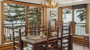 A dining area with a wooden table and chairs, a chandelier, and large windows with a view of trees and a snowy landscape. - On the Slopes, Recent Upgrades, Stonebridge Condo #935, Steps to Base Village (Snowmass Village)